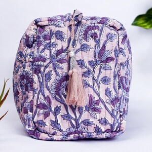 May include: A quilted cosmetic bag with a floral pattern in shades of purple and lavender. The bag has a drawstring closure with a tassel and bead embellishment. The bag is approximately 6 inches wide and 5 inches tall.