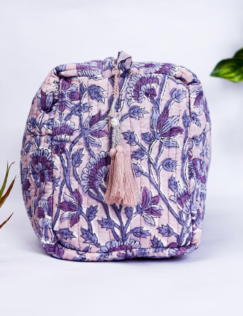 May include: A quilted cosmetic bag with a floral pattern in shades of purple and lavender. The bag has a drawstring closure with a tassel and bead embellishment. The bag is approximately 6 inches wide and 5 inches tall.