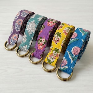 May include: Five colorful fabric belts with floral patterns and brass D-rings. The belts are in shades of purple, teal, green, yellow, and blue. Each belt has a unique floral design, offering a variety of styles.
