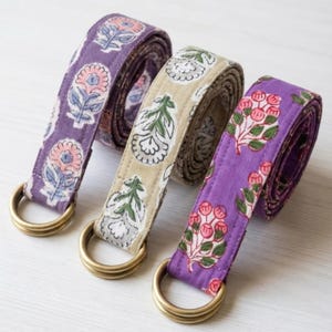 May include: Three fabric belts with floral patterns and brass-colored D-rings. The belts are in shades of purple and tan, with pink, green, and white floral designs. The belts are rolled up, showcasing the repeating patterns.