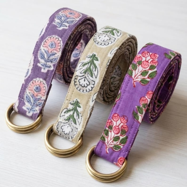 May include: Three fabric belts with floral patterns and brass-colored D-rings. The belts are in shades of purple and tan, with pink, green, and white floral designs. The belts are rolled up, showcasing the repeating patterns.