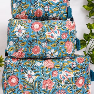 May include: Three stacked, rectangular cosmetic bags with a vibrant floral pattern. The bags feature a teal background with pink, white, and yellow flowers and green leaves. Each bag has a teal tassel zipper pull.