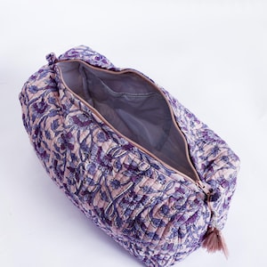 May include: Open, quilted travel bag with a floral pattern in shades of purple and pink. The bag has a zipper closure and a tassel. The interior is a solid gray color.