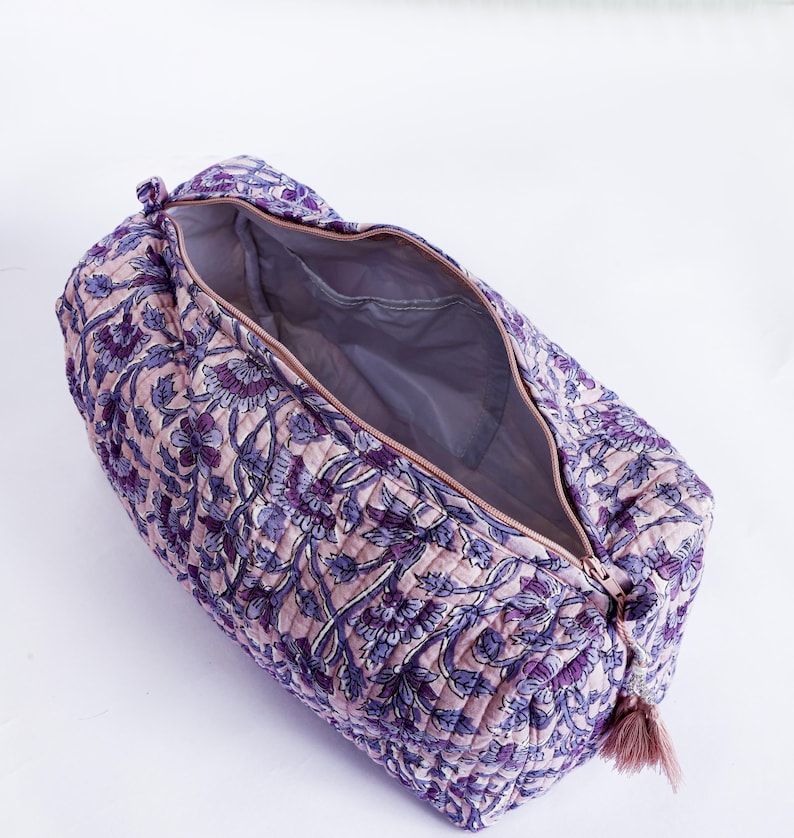 May include: Open, quilted travel bag with a floral pattern in shades of purple and pink. The bag has a zipper closure and a tassel. The interior is a solid gray color.