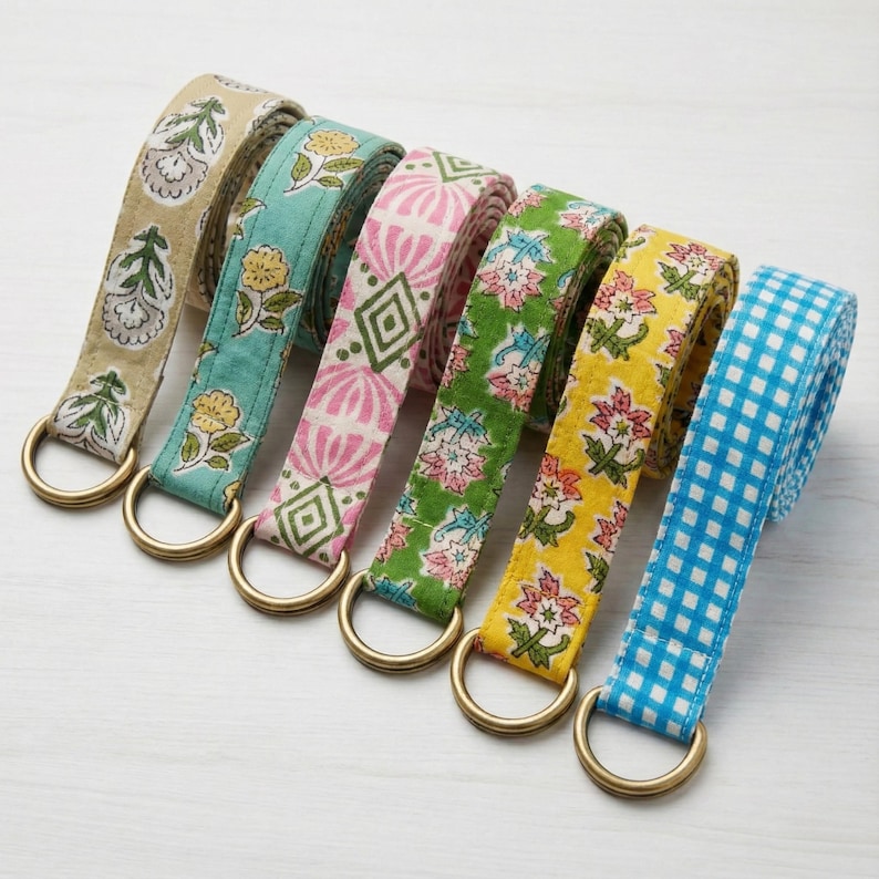 May include: A collection of seven fabric belts with brass-colored D-rings. The belts feature various patterns, including floral and geometric designs, in colors like beige, teal, pink, green, yellow, and blue. The belts are arranged in a row.