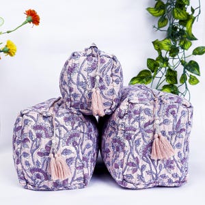 May include: Three quilted cosmetic bags with a floral pattern in shades of purple and white. Each bag has a matching tassel zipper pull. The bags are arranged in a stacked formation.