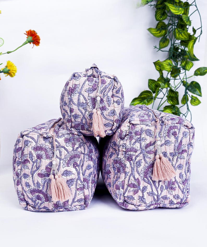 May include: Three quilted cosmetic bags with a floral pattern in shades of purple and white. Each bag has a matching tassel zipper pull. The bags are arranged in a stacked formation.