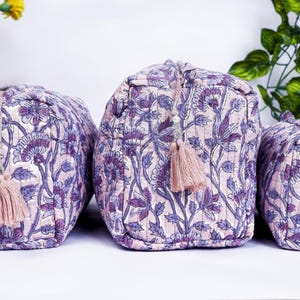 May include: Set of three quilted cosmetic bags in varying sizes. The bags feature a floral pattern in shades of purple and white, with matching pink tassels. The bags are designed for storing makeup and toiletries.