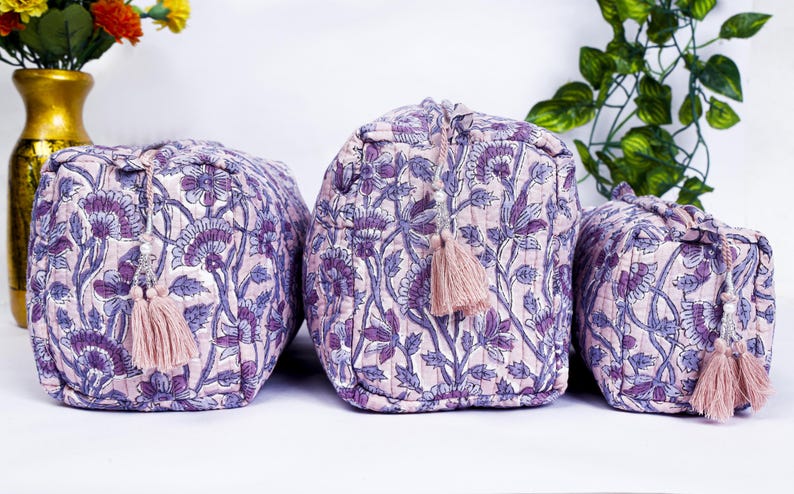 May include: Set of three quilted cosmetic bags in varying sizes. The bags feature a floral pattern in shades of purple and white, with matching pink tassels. The bags are designed for storing makeup and toiletries.