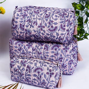 May include: Three quilted cosmetic bags in varying sizes, stacked. The bags feature a floral pattern in shades of purple and blue on a light pink background. Each bag has a matching tassel.