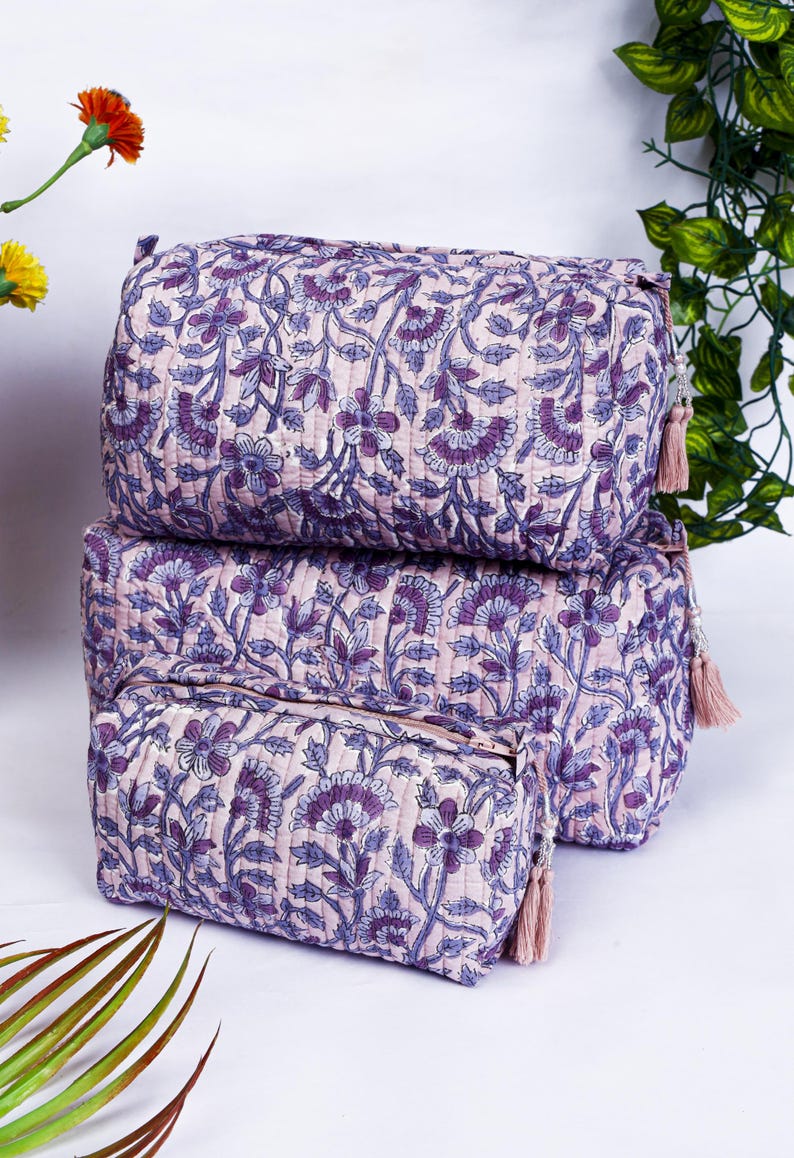 May include: Three quilted cosmetic bags in varying sizes, stacked. The bags feature a floral pattern in shades of purple and blue on a light pink background. Each bag has a matching tassel.