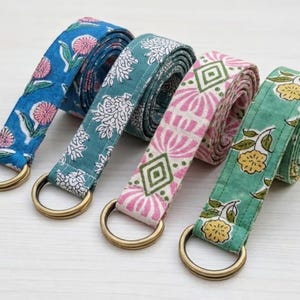 May include: Four colorful yoga straps with floral patterns. The straps are blue, teal, pink, and green, each with a different design. They have gold-colored metal D-rings. The straps are made of fabric and are rolled up.