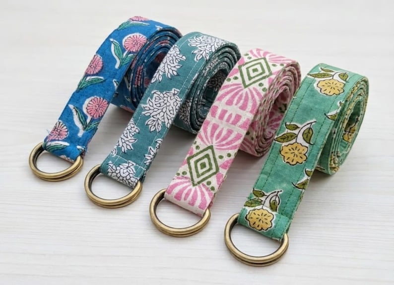 May include: Four colorful yoga straps with floral patterns. The straps are blue, teal, pink, and green, each with a different design. They have gold-colored metal D-rings. The straps are made of fabric and are rolled up.