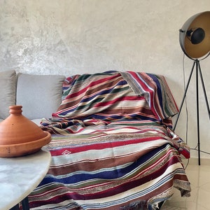 May include: A striped throw blanket in shades of red, blue, green, brown, and white is draped over a light gray couch. The blanket has a frayed edge and is made of a woven fabric.