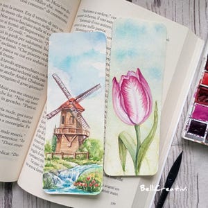 May include: Two watercolor bookmarks. One features a windmill scene with a waterfall and flowers. The other shows a pink tulip with green leaves. The bookmarks are on a book, with a watercolor palette and a pen nearby.