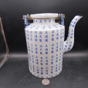 May include: A white and blue ceramic pitcher with a curved spout and two handles. The pitcher is decorated with blue Chinese characters. The handles are gold and blue. A coin is at the base of the pitcher.