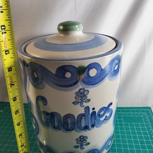 May include: A ceramic cookie jar with a lid, painted with blue swirls and the word "Goodies". The jar is approximately 9 inches tall, with a green knob on the lid. The jar is on a green surface.
