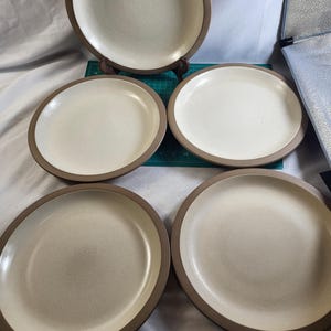 May include: A set of six beige ceramic plates with a speckled finish and a brown rim. The plates are round and vary in size, with one plate displayed on a wooden stand. The plates are suitable for serving meals.