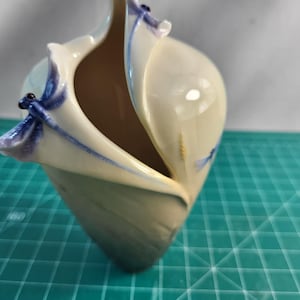 May include: A cream-colored ceramic vase with a unique split-top opening. The vase features delicate, hand-painted purple and blue dragonfly motifs perched on the edges of the rim, with a smooth, glossy finish and a tapered, pointed base.