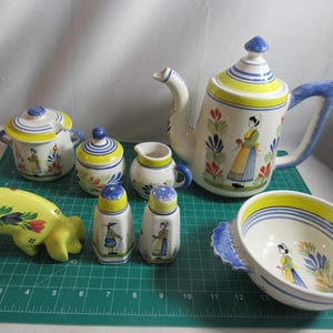 May include: A collection of ceramic kitchenware with a Dutch theme. Includes a teapot, sugar bowl, creamer, salt and pepper shakers, a serving bowl, and a decorative pig. The pieces are white with blue, yellow, and green accents.