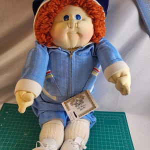 May include: A Cabbage Patch Kids doll with orange yarn hair, blue eyes, and a soft fabric face. The doll wears a blue corduroy zip-up jacket with white ribbed cuffs, matching blue pants, white socks, and white shoes, topped with a blue baseball cap.