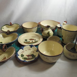 May include: A collection of hand-painted ceramic bowls, dishes, and a small pitcher. The pieces are cream-colored with blue, green, and brown accents. Some have text, and illustrations of houses and trees. A spoon rests in a small bowl.