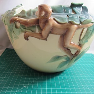 May include: Cream-colored ceramic bowl with a monkey climbing on the side. The monkey is brown with a light-colored face and hands. Green leaves and accents are around the rim of the bowl. The bowl is sitting on a green cutting mat.