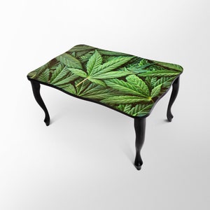 Op de afbeelding: Een zwarte salontafel met een groene marihuanabladprint op de bovenkant. De tafel heeft vier poten met een gebogen ontwerp.
