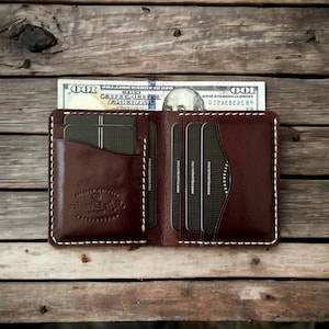 May include: Open brown leather wallet with white stitching, holding several cards and a one hundred dollar bill. The wallet is on a wooden surface. The wallet has a logo on the inside.