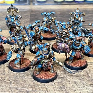 May include: A collection of miniature figurines, primarily in shades of blue and gold, with intricate armor and weaponry. The figures are posed on circular bases with a textured, brown surface, suggesting a tabletop game or hobby theme.
