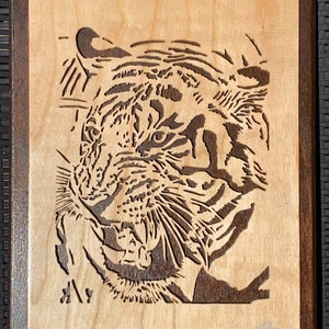 May include: A close-up of a wood-carved tiger head, with intricate details of the fur and facial features. The tiger's mouth is slightly open, showing its teeth. The carving is mounted on a wooden plaque with a dark brown frame.