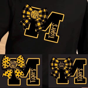 May include: A black long-sleeve shirt featuring a large letter M with a yellow outline and the word Tigers written vertically. The design includes a yellow and black plaid bow with the text Miss Our in the centre, plus two variations.