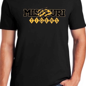 Puede incluir: Una camiseta negra de manga corta con el texto MISSOURI en letras negras con borde amarillo y un gráfico de marcas de garras. Debajo, la palabra TIGERS está impresa en negro dentro de seis diamantes amarillos en una fila horizontal.