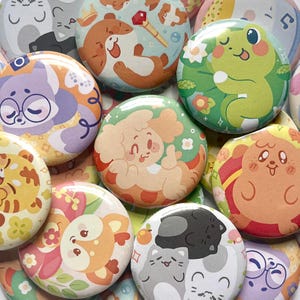 May include: A selection of colourful, round pinback buttons displaying cartoon animal illustrations. Designs include a frog, dog, cat, and other creatures, with floral and patterned backgrounds. The buttons are in shades of green, orange, purple, and yellow.