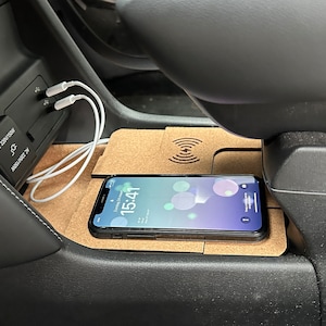 Peut inclure: Gros plan sur la console centrale d'une voiture avec un chargeur sans fil et un téléphone dessus. Le téléphone est en charge et l'écran affiche l'heure 15:41. Il y a également deux cordons blancs branchés sur la console.