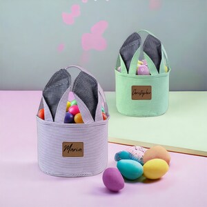 May include: Two Easter baskets with bunny ears, one green and one white with black stripes. The green basket has the name "Christopher" on it and the white basket has the name "Maria" on it. Both baskets are filled with colorful plastic Easter eggs.