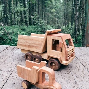 May include: Two wooden toy trucks, one a dump truck and the other a smaller pickup truck, are on a wooden surface. The trucks are made of natural wood and have a rustic look.