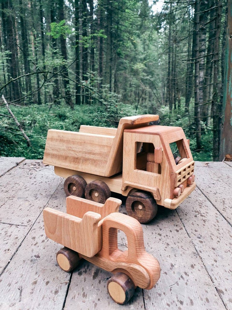May include: Two wooden toy trucks, one a dump truck and the other a smaller pickup truck, are on a wooden surface. The trucks are made of natural wood and have a rustic look.