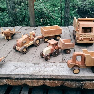 May include: A collection of wooden toy vehicles, including a tractor, a dump truck, a pickup truck, a plane, and a flatbed truck. The toys are all made of natural wood and have a rustic, handmade look.