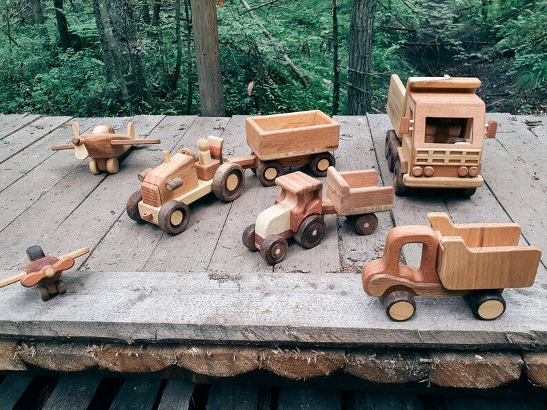 May include: A collection of wooden toy vehicles, including a tractor, a dump truck, a pickup truck, a plane, and a flatbed truck. The toys are all made of natural wood and have a rustic, handmade look.