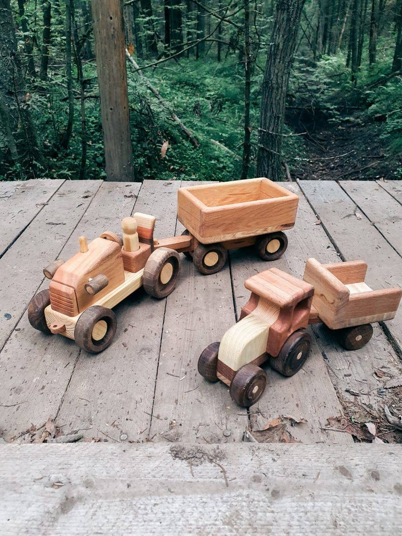 May include: Three wooden toy tractors with trailers. The tractors are brown and natural wood with black wheels. The trailers are brown wood with no wheels. The toys are on a wooden surface.