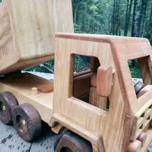 May include: A wooden toy dump truck with a brown bed and a light brown cab. The truck has four wheels and a detailed design. The truck is sitting on a wooden surface.