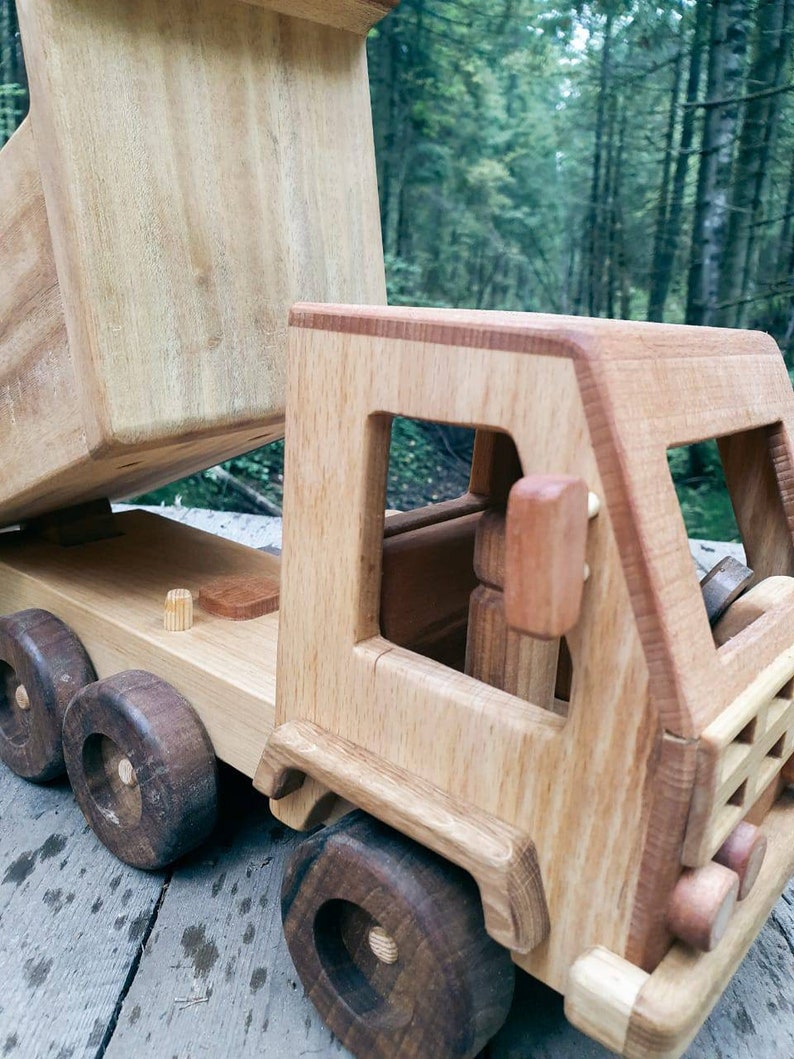 May include: A wooden toy dump truck with a brown bed and a light brown cab. The truck has four wheels and a detailed design. The truck is sitting on a wooden surface.