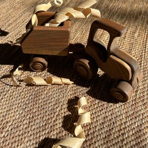 May include: A wooden toy truck with a trailer, both made of dark brown wood. The trailer is filled with wood shavings. The truck and trailer are sitting on a brown woven mat.