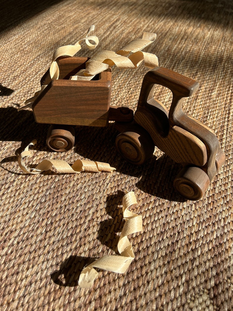 May include: A wooden toy truck with a trailer, both made of dark brown wood. The trailer is filled with wood shavings. The truck and trailer are sitting on a brown woven mat.
