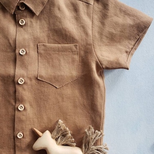 May include: A brown short-sleeved shirt with a button-down design and a chest pocket. The shirt is made of a textured fabric. A wooden unicorn toy with twine mane and tail is placed below the shirt. The shirt has wooden buttons.