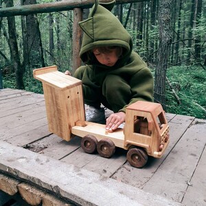 May include: A child wearing a green hooded sweatshirt plays with a wooden toy truck in a forest setting. The truck has a flatbed and a separate wooden cab. The truck is made of natural wood with no paint.