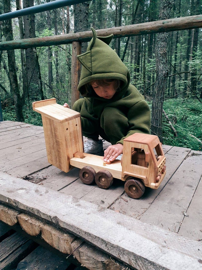 May include: A child wearing a green hooded sweatshirt plays with a wooden toy truck in a forest setting. The truck has a flatbed and a separate wooden cab. The truck is made of natural wood with no paint.