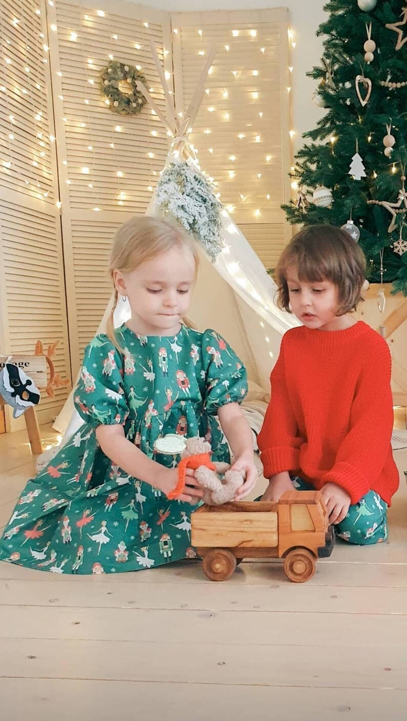 May include: Two young girls play with a wooden toy truck and a stuffed animal. The girl on the left is wearing a green dress with a Nutcracker pattern. The girl on the right is wearing a red sweater and green pants. The truck is brown and has a wooden bed. The stuffed animal is brown and has a red scarf.