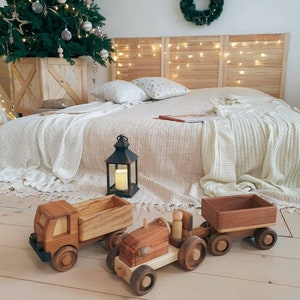 May include: A wooden toy tractor with a trailer, both painted in a natural wood color. The tractor has a small wooden figure in the driver's seat. The toys are on a light wood floor.
